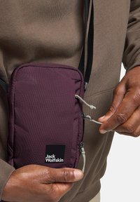 Paarse nylon tas met rits, verstelbare schouderriem en logo. Hand die aan de rits trekt, gedragen over een beige sweatshirt. Gestructureerde stof.