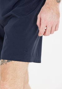 Tummansininen lyhyt housut, jotka on valmistettu kevyestä kankaasta ja joissa on joustava vyötärö. Vasemmalla puolella on pieni brodeerattu logo.