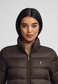 Brun puffjacka med hög krage, quiltat mönster, framdragkedja och liten logotypdetalj på bröstet. Bärs över en texturerad beige tröja.