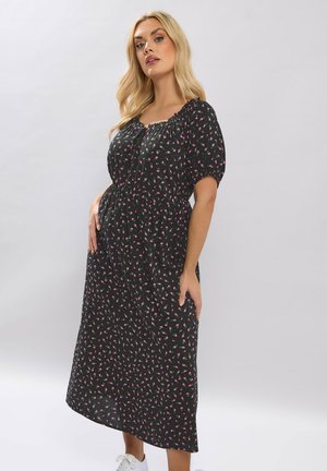 Lyshåret kvinde iført sort blomsterprintet midi-kjole med pufærmer, poserer foran en ensartet lysegrå baggrund.