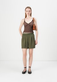 Hnedý smocked top, zelené plisované šortky, čierne šnurovacie baleríny a malá hnedá kabelka na rameno. Jednoduché pozadie zvýrazňuje oblečenie.