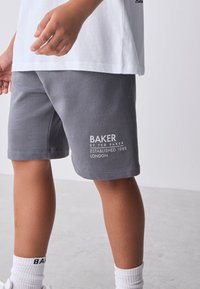 Asmuo, dėvintis pilkus šortus su užrašu „Baker by Ted Baker Established 1988 London“ ir baltą marškinėlį, stovintis ant šviesios grindų dangos.