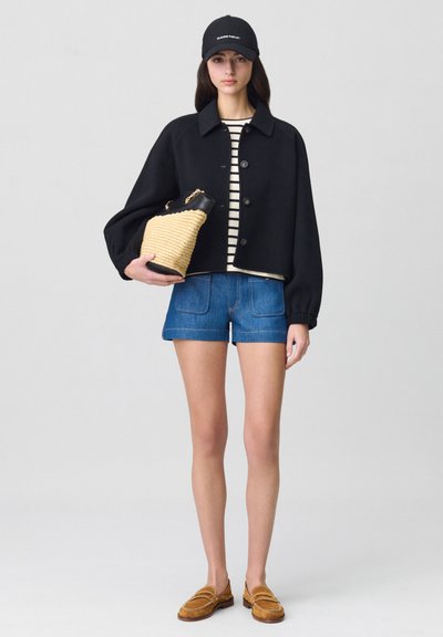 Veste cropped noire avec des boutons, chemise rayée blanche et noire, shorts en denim bleu, mocassins jaune-brun et sac en paille texturé.