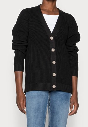 Personne portant un cardigan noir boutonné sur un haut blanc et un jean bleu, avec les manches légèrement remontées.