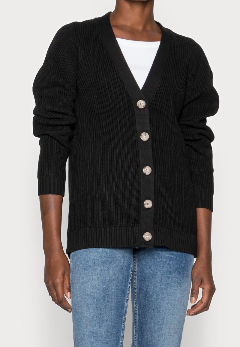 Personne portant un cardigan noir boutonné sur un haut blanc et un jean bleu, avec les manches légèrement remontées.