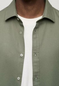 Camicia di cotone verde oliva con una finitura testurizzata. Caratterizzata da un design con bottoni, colletto e una T-shirt bianca indossata sotto.