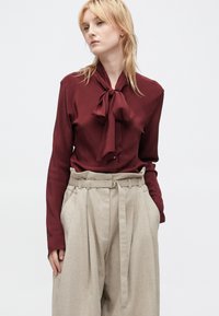 Bordeauxfarbene Bluse mit einer Schleifenkragen-Detail, langen Ärmeln und hellbraunen hochgeschnittenen Hosen mit Falten und einem breiten Gürtel. Glatte Textur.