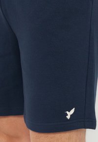 Κοντινό πλάνο σκούρων μπλε σορτς με μικρό λευκό κεντημένο λογότυπο κολιμπρί κοντά στο στρίφωμα στο αριστερό πόδι.