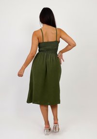 Vestido midi verde oliva con tirantes finos, cuerpo fruncido y falda fluida. Hecho de una tela suave con un acabado ligeramente texturizado. Tacones transparentes.