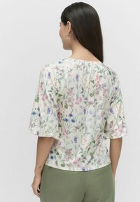 Blomsterbluse med korte puffærmer i kremfarve, med flerfarvede blomstermønstre. Knappedetalje ved den bageste halsudskæring, letvægtigt stof.