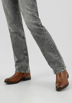 Benen in vervaagde grijze jeans en bruine enkellaarzen met lage hakken en gouden details op een eenvoudige lichte achtergrond.