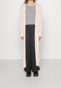 Jasnoróżowy kardigan z długim rękawem, otwarty z przodu, noszony na czarno-białą bluzkę w paski i szerokie czarne spodnie, zestawiony z czarnymi butami.