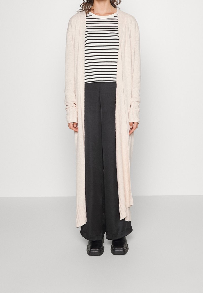 Jasnoróżowy kardigan z długim rękawem, otwarty z przodu, noszony na czarno-białą bluzkę w paski i szerokie czarne spodnie, zestawiony z czarnymi butami.