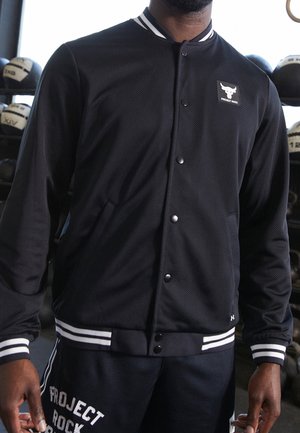 Czarna kurtka varsity z białymi paskowanymi mankietami i kołnierzem, logo Project Rock na piersi, noszona przez osobę na siłowni, z ułożonymi za nią piłkami lekarskimi.