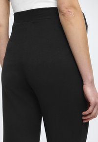 Schwarze, gerippte Hose mit hohem Bund, zeigt glatte Textur und nahtloses Design. Hand ruht am Bein und hebt die Passform des Stoffs hervor.