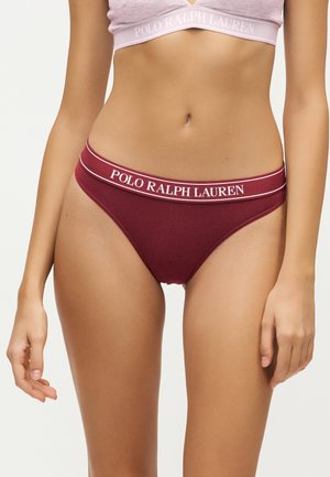 Жена, носеща бордово бельо Polo Ralph Lauren и светлорозов бюстие, показваща корема и ръцете срещу обикновен фон.