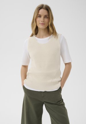 Femme aux cheveux châtain clair portant un gilet sans manches en maille crème sur une chemise blanche à manches courtes et un pantalon vert olive, les mains dans les poches.