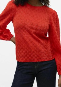 Rød tekstureret bluse med rund hals og puflede lange ærmer, båret med mørkeblå jeans. Stoffet har et mønster af små, hævede prikker.