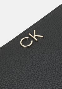 Svart läderplånbok med en texturerad yta och en framträdande guld "CK"-logotyp. Designen är strömlinjeformad med en slät finish.