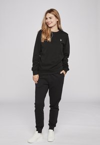 Zwarte sweatshirt en broek van een katoenmix. De sweatshirt heeft ribbels aan de mouwen en een klein logo op de borst. Het model draagt witte sneakers.