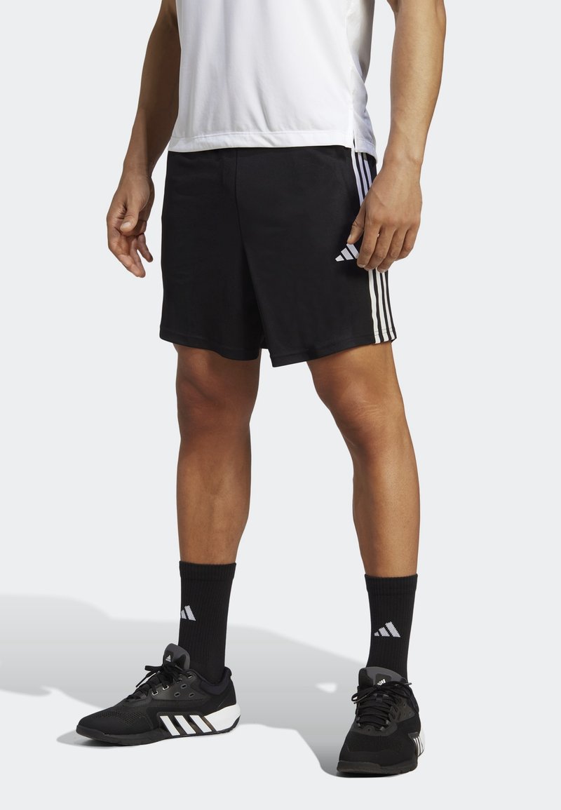 Pantalones cortos deportivos negros con tres rayas blancas en los lados, combinados con calcetas negras y zapatos con un toque blanco. Camiseta sin mangas blanca.