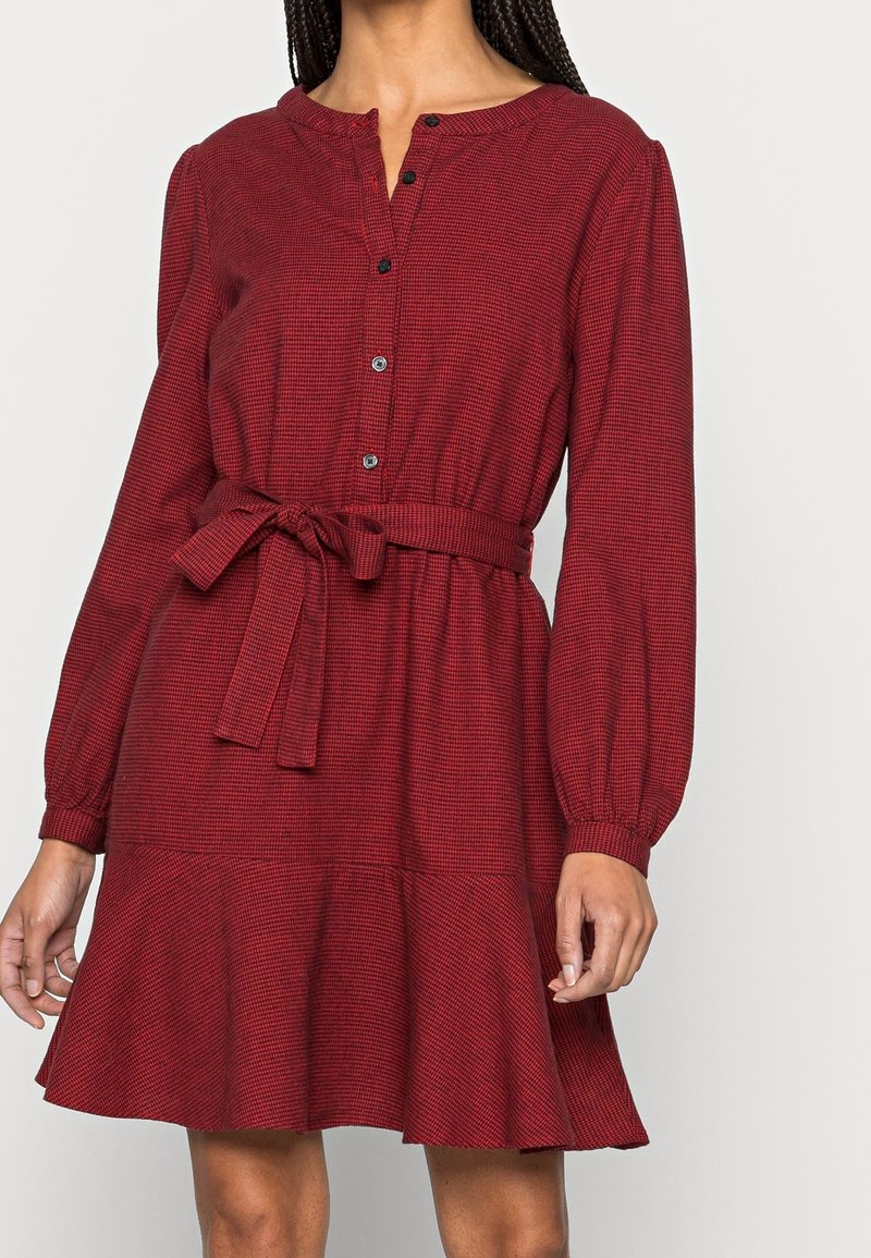 Femme portant une robe longue à manches longues rouge ceinturée, avec un petit motif à carreaux et un col boutonné, se tenant devant un fond uni.