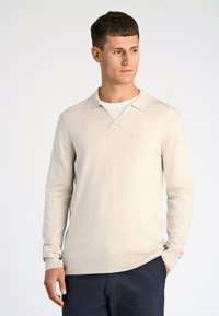 Ljusbeige stickad pullover med V-ringning och långa ärmar. Tyget verkar mjukt och strukturerat med en diskret logotyp på bröstet.