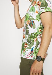 Camisa de manga corta con estampado tropical que presenta hojas verdes y flores coloridas, sobre un fondo blanco; combinada con pantalones verde oliva.