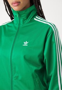 Zelená polyesterová bunda na zip s vysokým límcem, s bílými třípruhovými akcenty na rukávech a malým bílým logem Adidas na hrudi.