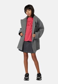 Niña pequeña con un abrigo gris oversized, camiseta gráfica rosa, falda de mezclilla oscura y zapatillas negras, de pie contra un fondo blanco.