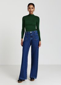 Grünes, geripptes Langarmoberteil, kombiniert mit hoch taillierten, weiten blauen Jeans. Auffällige Details sind die vorderen Taschen und der Knopfverschluss.