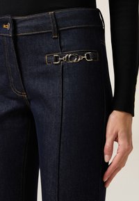 Los jeans de mezclilla oscura presentan costuras amarillas, un bolsillo lateral con accents de metal plateado y un diseño ajustado, destacando una silueta limpia y entallada.