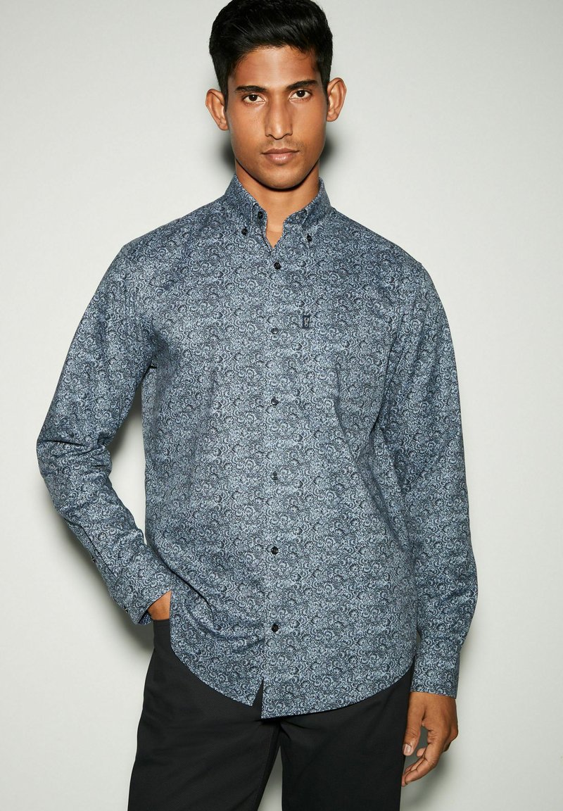 Camisetta a maniche lunghe di colore blu navy con un motivo paisley, dotata di colletto a camicia e bottoni neri. Il tessuto appare liscio e leggero.