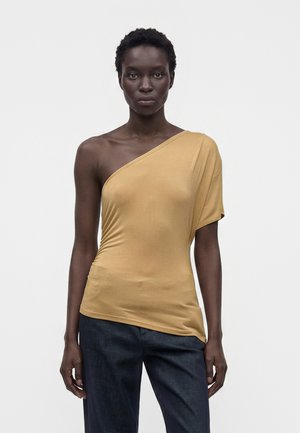 Femme portant un haut épaule dénudée jaune moutarde à manches courtes et un pantalon bleu foncé, debout devant un fond blanc uni.