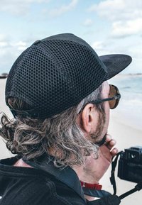 Black cap with mesh design, featuring a flat brim. Long, wavy gray hair visible, wearing sunglasses. A camera held in one hand.