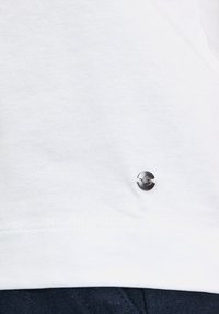 T-shirt en coton blanc avec un accent de logo rond argenté près de l'ourlet. Texture douce avec un design épuré et simple.