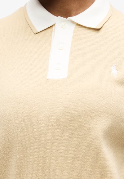 Μπεζ πλεκτό πόλο πουκάμισο με λευκό γιακά και πατιλέτα, κουμπωμένο, με μικρό κεντητό λογότυπο στη δεξιά πλευρά του στήθους.