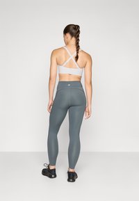Soutien-gorge de sport gris clair avec des bretelles croisées, associé à un legging taille haute gris foncé et des chaussures de sport noires. Tissu lisse et extensible.