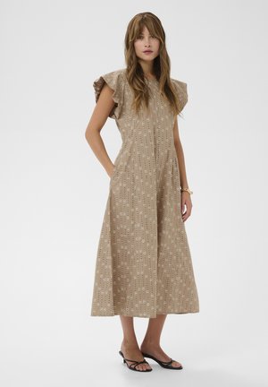 Femme en robe midi beige à motif floral, manches à volants, sandales noires à talons, debout devant un fond blanc avec les mains dans les poches.