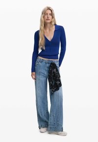 Sinivihreä ribattu pitkähihainen paita, jossa on kaulus, yhdistettynä leveälahkeisiin vaaleisiin farkkuihin. Asusteena musta kuvioitu huivi solmittuna vyötärölle.