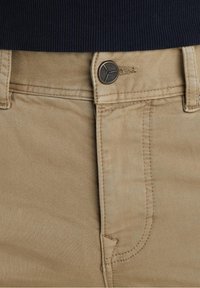 Pantalones de algodón beige con una textura suave, que cuentan con una bragueta con cremallera y un botón metálico redondo con grabado de la marca sobre el bolsillo delantero.