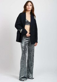 Giovane donna che indossa pantaloni in velluto schiacciato argento e un bralette abbinato con un blazer oversize nero caratterizzato da dettagli a fiocco in strass.