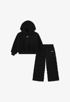 Felpa nera con cappuccio Nike in tessuto soffice e pantaloni abbinati a gamba larga con logo swoosh bianco sul petto e sulla coscia sinistra su sfondo bianco.