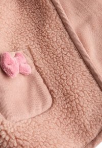 Tissu polaire rose doux avec un motif texturé, avec une poche côtelée ornée d'un petit nœud rose en peluche.