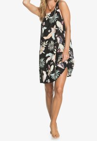 Femme portant une robe noire sans manches à imprimé floral tropical coloré, debout pieds nus, levant l'ourlet de la robe d'une main.