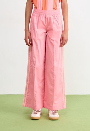 Personne portant un pantalon de survêtement rose ample avec des bandes latérales et des baskets blanches sur un fond clair uni.