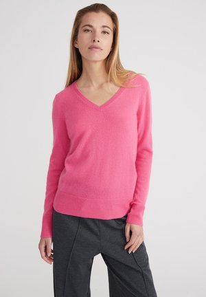 Sweter z dekoltem w serek, z długimi rękawami w jaskrawym różowym kolorze, wykonany z miękkiego materiału z prążkowanymi ściągaczami i dołem. Połączony z szarymi, teksturowanymi spodniami.