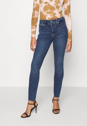 Žena nosící tmavě modré džíny s vysokým pasem a černé sandály na vysokém podpatku se stahovacími pásky, doplněné dlouhým hnědo-bílým topem s batikovaným vzorem.