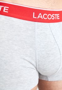 Γκρίζα βαμβακερή σλιπ με κόκκινη ελαστική ζώνη που φέρει λευκό κείμενο "LACOSTE". Λείο υλικό και εφαρμοστή σχεδίαση.
