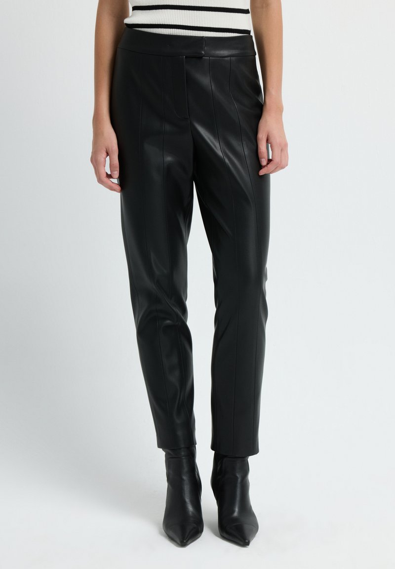 Zwarte faux leren broek met een toelopende pasvorm, verticale stiksel details en een gladde textuur, gecombineerd met puntige zwarte enkellaarzen.